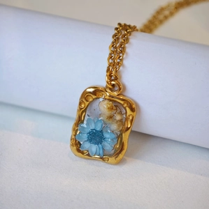 Dettaglio del ciondolo rettangolare dorato con margherite blu essiccate e fiori
