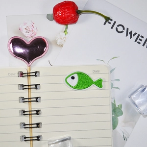 Mini green fish embroidery patch and pink heart charm on a white notebook page