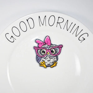 Cartoon owl embroidery patch with pink glasses and bow on a white ceramic plate