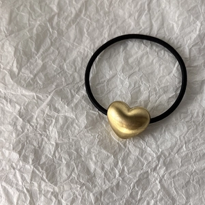 Detail of a matte gold heart-shaped charm on a high-elastic black hair tie over textured paper