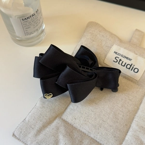 Detail of a black satin layered bow hair clip with a gold heart charm on a beige texture