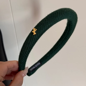 Dettaglio cerchietto in maglia verde scuro con ciondolo a uccello dorato ed etichetta Hand Made