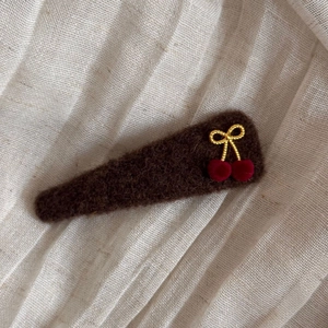 Close-up of brown felt hair clip featuring a gold bow and red plush cherries on white linen