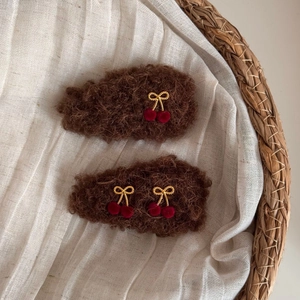 Brown plush bow hair clips with cherry charms on a minimalist woven basket background