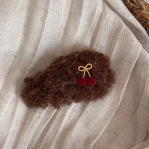 Detail of plush brown hair clip with golden bow and red cherry charms on linen