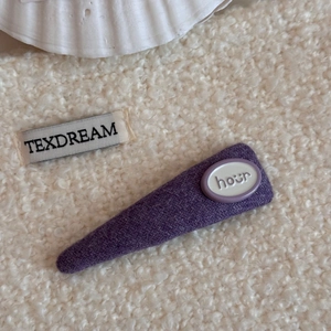 Detalle de pinza para el pelo en tela violeta con etiqueta blanca hour sobre fondo de felpa