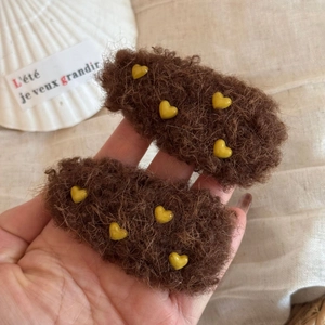 Braune Plüsch-Haarspangen mit gelben Herzen über einer weißen Muschelschale