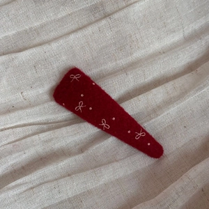 Dettaglio molletta in feltro rosso con fiocco ricamato su sfondo di lino testurizzato