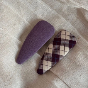 Detail view of two fabric BB hair clips in solid purple and plaid patterns on beige linen