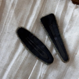 Detail view of two black felt-covered BB hair clips on a soft white linen background