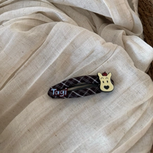 Close-up of brown plaid hair clip featuring a beige cartoon bear and red Tagi logo tag