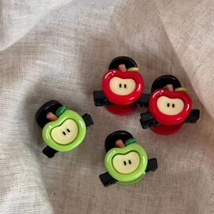 Detailed view of red and green apple-shaped mini hair clips on natural linen fabric