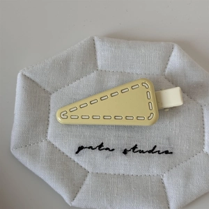 Detail of a creamy yellow triangular hair clip with embossed white stitching on a linen base