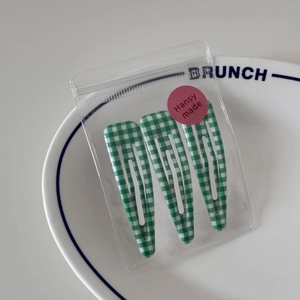 Detailed view of three green gingham BB hair clips in a transparent bag on a white plate