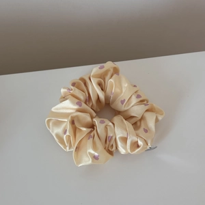 Close-up of beige satin scrunchie with purple heart patterns and a silver brand charm
