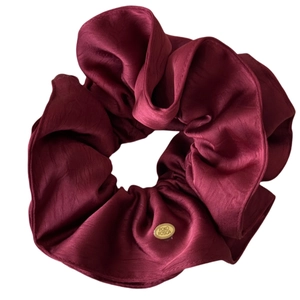 Elegant wine red satin scrunchie with golden oval logo tag and rich pleated texture