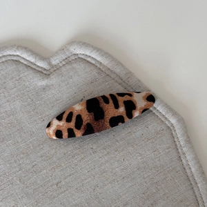 Detail showing the oval leopard print hair clip with fabric texture on a beige mat