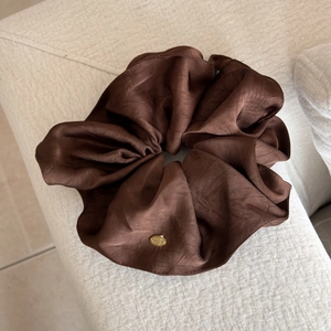 Detail eines braunen Satin-Haargummis mit einer goldenen ovalen Logo-Plakette
