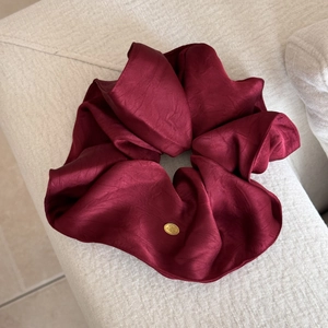 Detailaufnahme von burgunderfarbenem Satin-Scrunchie mit einer goldenen Logo-Metallplatte