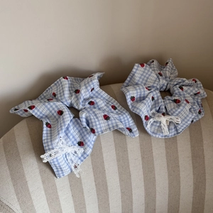 Detail view of two blue plaid strawberry scrunchies with white lace bows on fabric