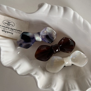 Close-up of three acetate bow hair clips in purple, brown, and pearl white on a shell tray