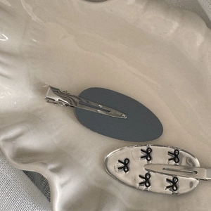 Detail view of oval hair clips featuring matte grey and silver bow designs on a ceramic tray