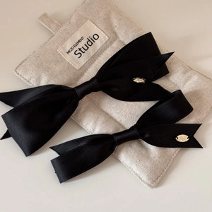 Detail of two black satin bow hair clips with gold metal logo on beige fabric