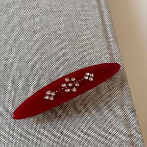 Close-up of a red oval hair clip with sparkling rhinestone flower motifs on gray fabric