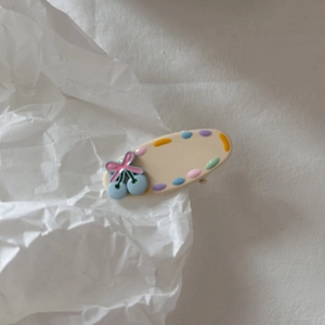 Dettaglio molletta per capelli crema con ciliegia blu e fiocco rosa su carta