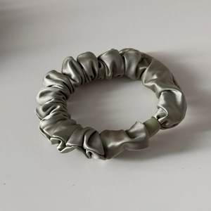 Nahaufnahme eines silbergrauen Satin-Scrunchies mit Falten und grünem Deko-Verschluss