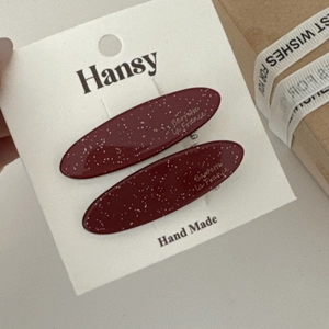 Dettaglio di fermagli per capelli in acetato rosso vino con glitter e logo dorato