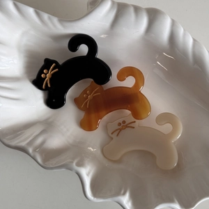 Three acetate cat-shaped hair clips in black, amber, and beige on a shell tray