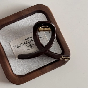 Detailed close-up of dark brown French hair claw clip with golden logo in a wooden tray