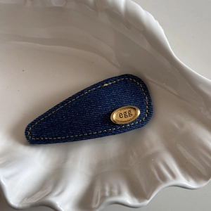 Detail of denim hair clip with gold egg logo plate on a white shell plate