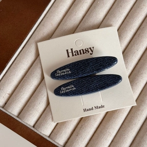 Detailed view of two denim texture hair clips with silver letter labels on display card