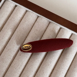 Close-up of a burgundy fabric bow hair clip with a gold metal Korea logo plate