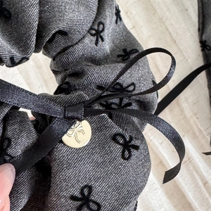 Macro shot of gray denim scrunchie featuring black bows and a gold logo pendant