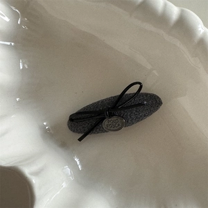 Détail d'une pince à cheveux en tissu gris avec nœud noir et étiquette métallique ronde