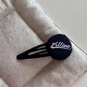 Close-up of black fabric BB clip with white Alline embroidery on a beige texture