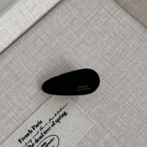 Detail of a black teardrop hair clip with golden France Handmade text on beige fabric