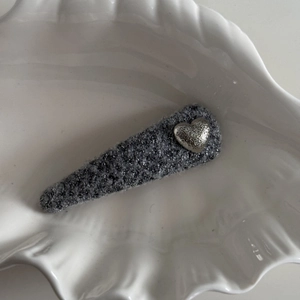 Detail of gray tweed BB hair clip with silver heart charm on white shell prop