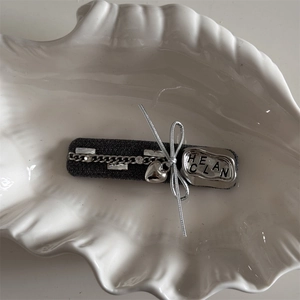 Detail of grey fabric hair clip with metal chain, heart charm and HELAN seal on shell tray