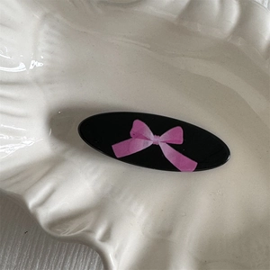 Close-up of black oval hair clip with pink bow pattern on a white shell-shaped plate
