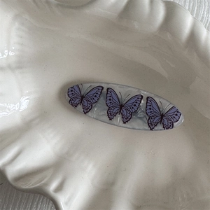 Detail view of oval acetate hair clip with elegant purple butterfly patterns on a white tray