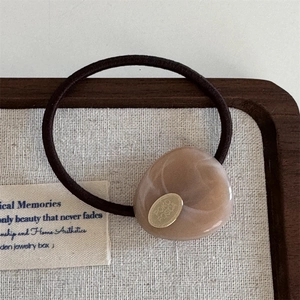 Close-up of brown elastic hair tie featuring resin pebble charm and gold oval metal label