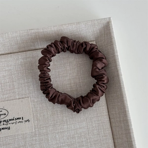 Detailed shot of dark brown silk satin scrunchie with soft pleats in a beige woven tray