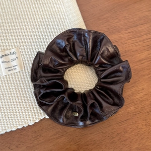 Detail of dark brown leather scrunchie with shiny finish and a round gold metal logo charm