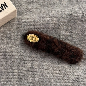 Close-up of a brown fuzzy hair clip with an oval gold brand tag on a grey fabric