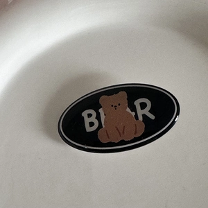 Détail d'une pince à cheveux ovale noire en résine avec un ours brun et l'inscription BEAR