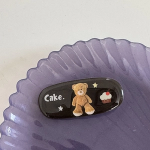Détail d'une pince à cheveux noire avec ours en peluche et cupcake sur un plateau